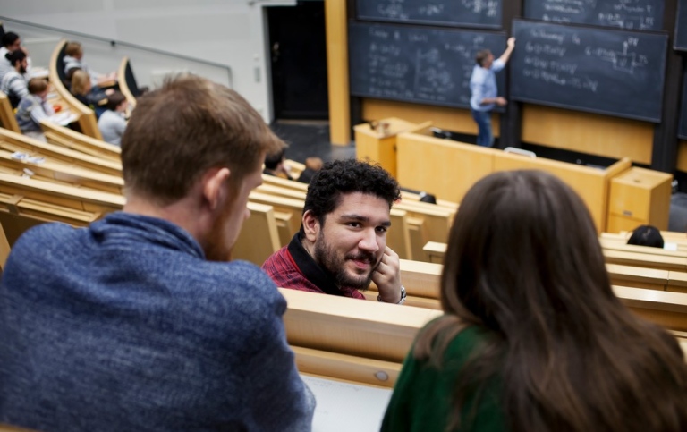 Conversation in auditorium, someone writing on blackboard in the background.